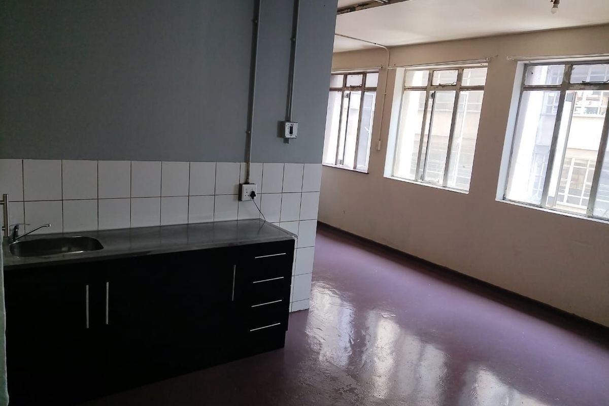 An open-plan living and kitchen area featuring a basic kitchenette with a stainless steel sink, tiled splashback, and large windows providing natural light.