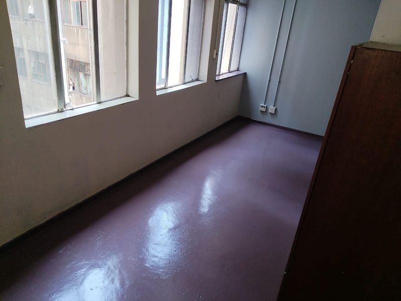 An empty bedroom featuring painted concrete flooring, large windows with aluminium frames, and exposed electrical conduit along the wall.
