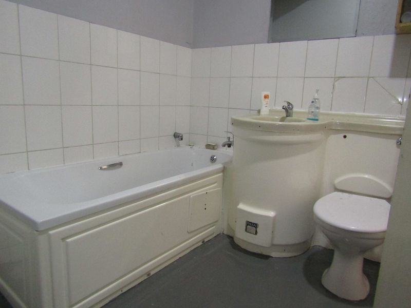 A dated bathroom featuring a standard bathtub, pedestal washbasin, and toilet with white wall tiling and grey flooring.