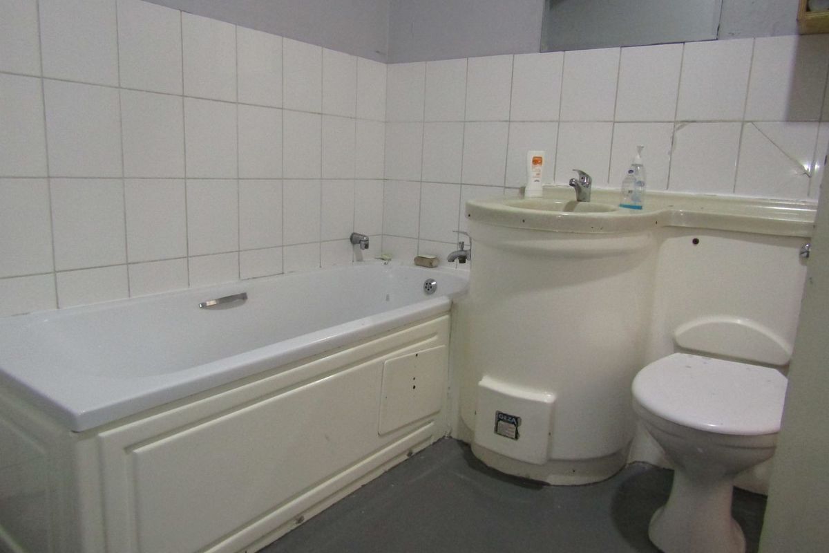 A dated bathroom featuring a standard bathtub, pedestal washbasin, and toilet with white wall tiling and grey flooring.