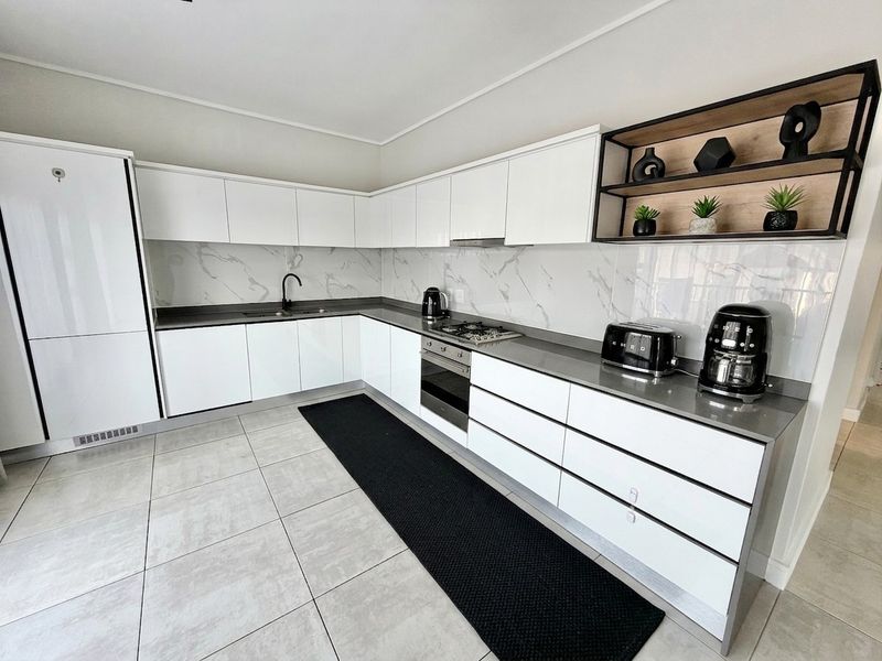 Modern kitchen featuring sleek white cabinetry, stone countertops, marble-look splashback, integrated oven, gas hob, and open shelving.