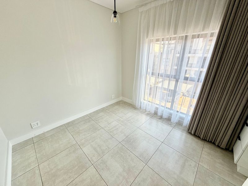 A bright, empty bedroom featuring modern large-format floor tiles, a large window with sheer curtains, and a minimalist pendant light fixture.