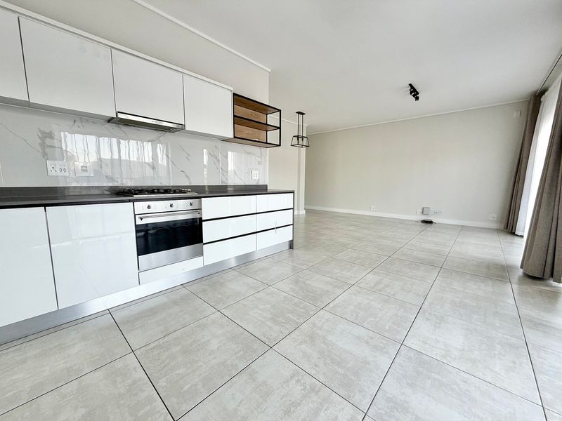 Modern open-plan kitchen and living area featuring sleek white cabinetry, integrated appliances, marble-look splashback, and neutral tiled flooring throughout.