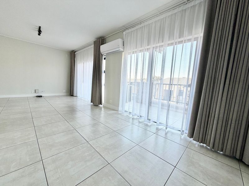 Spacious, light-filled living area featuring large format floor tiles, split-system air conditioning, and sliding glass doors leading to a balcony.