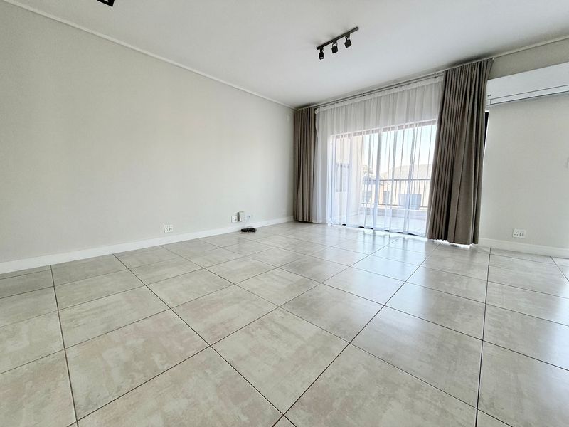 A bright, empty living room featuring large ceramic floor tiles, track lighting, and a sliding glass door leading to a balcony.