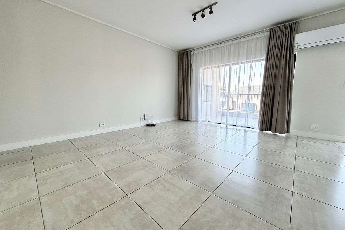 A bright, empty living room featuring large ceramic floor tiles, track lighting, and a sliding glass door leading to a balcony.