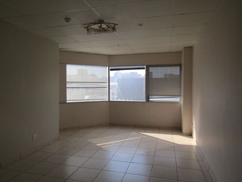 An empty living area featuring tiled flooring, a drop ceiling, and a bay window with venetian blinds offering an urban view.