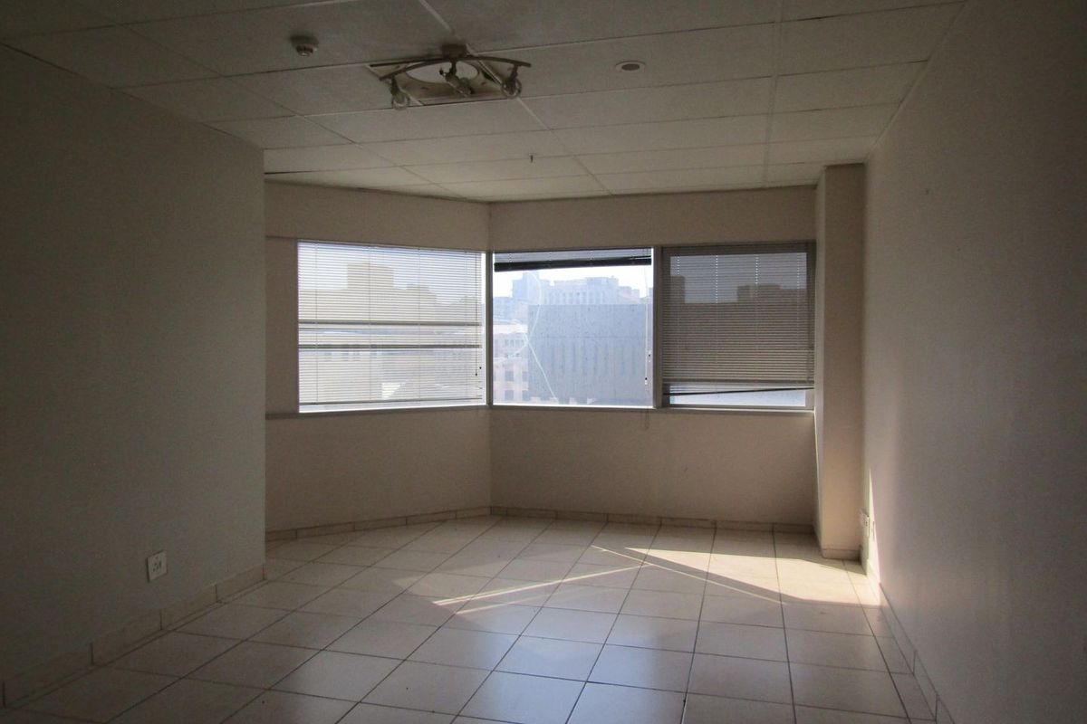 An empty living area featuring tiled flooring, a drop ceiling, and a bay window with venetian blinds offering an urban view.