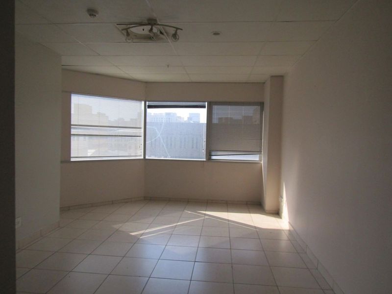 An empty, open-plan living area featuring tiled flooring, a suspended ceiling, and large windows with venetian blinds offering natural light.