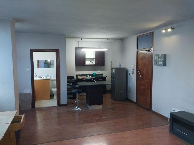 Open plan living area featuring a compact kitchen with dark cabinetry and laminate flooring, with direct access to an adjacent bathroom.