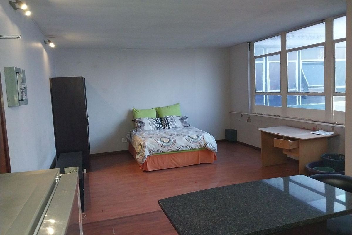 Open-plan studio apartment featuring laminate flooring, large windows providing natural light, and track lighting. The space requires cosmetic updates to improve its overall condition.