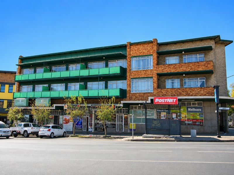 A multi-story commercial building featuring ground-floor retail storefronts, upper-level balconies with green railings, and brick facade detailing along a street-facing frontage.