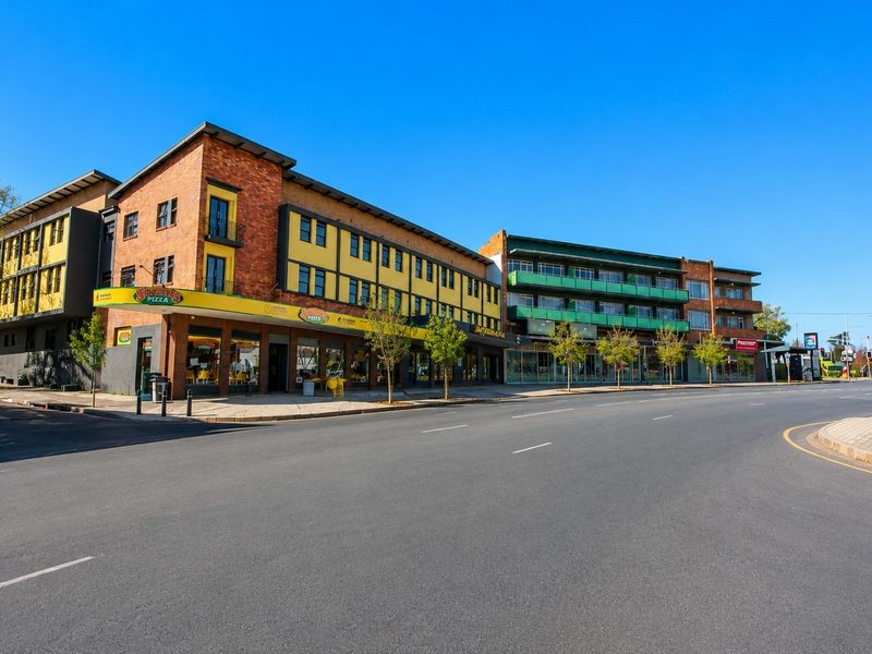 A street-level view of a multi-story commercial building featuring brick and yellow facade elements, ground-floor retail spaces, and upper-level balconies.