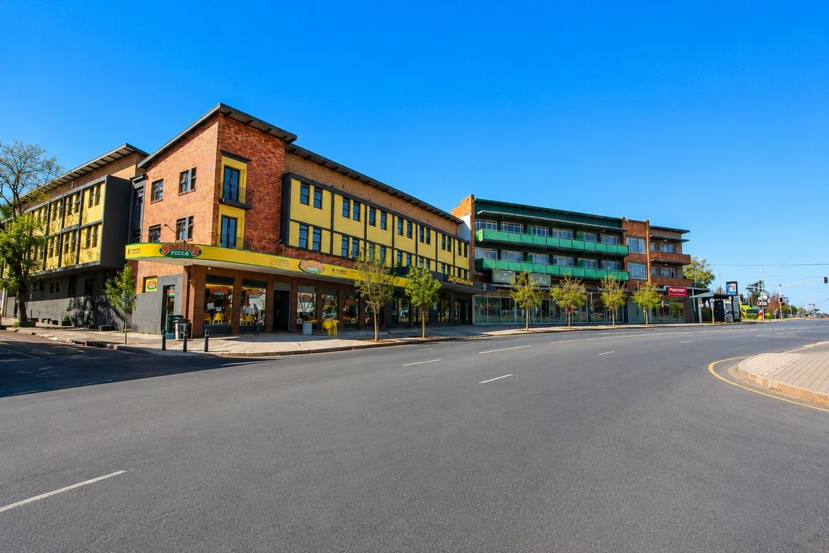 A street-level view of a multi-story commercial building featuring brick and yellow facade elements, ground-floor retail spaces, and upper-level balconies.