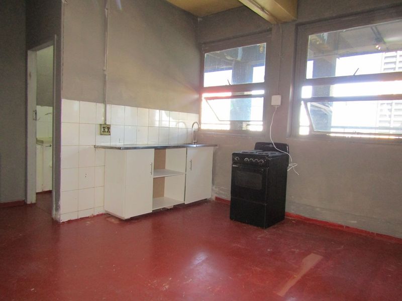 A basic, unfinished kitchen area featuring a small sink unit, tiled splashback, and a freestanding stove, requiring significant renovation and cosmetic updates.
