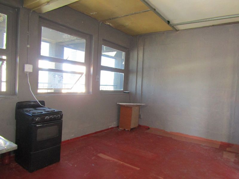A basic, empty kitchen space featuring a freestanding stove, exposed electrical conduit, and large windows, requiring significant renovation and cosmetic updates.