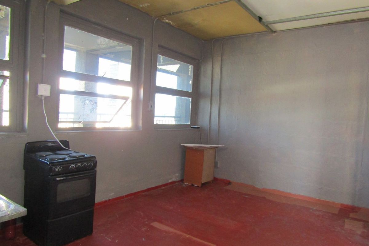 A basic, empty kitchen space featuring a freestanding stove, exposed electrical conduit, and large windows, requiring significant renovation and cosmetic updates.