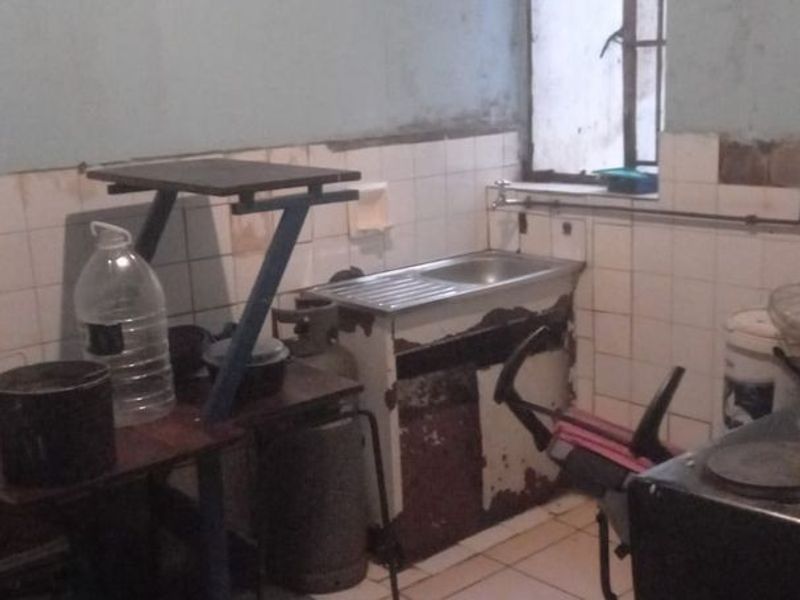 A dated kitchen area featuring a stainless steel sink, tiled splashback, and tiled flooring, currently requiring significant renovation and repair to wall and cabinet finishes.