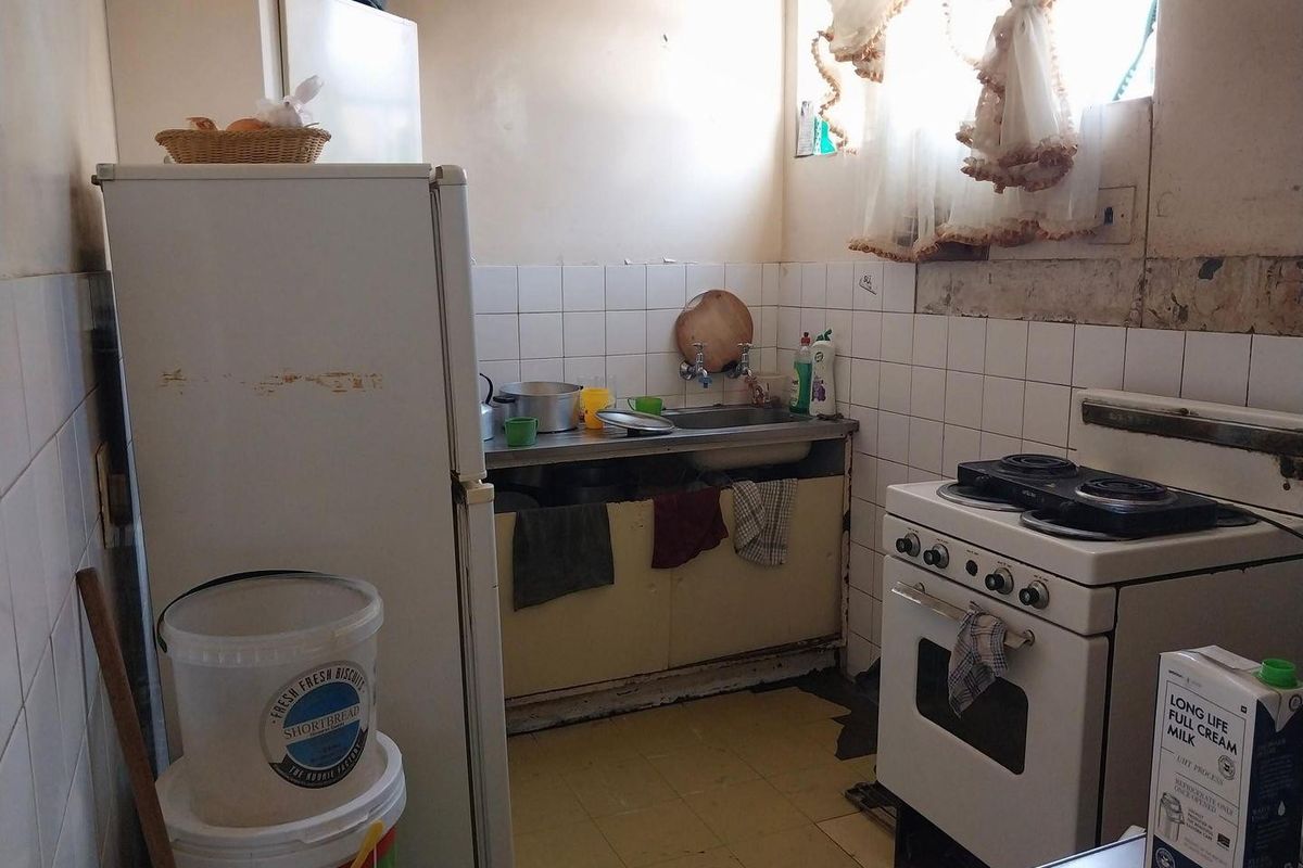 A compact, dated kitchen featuring a stainless steel sink, tiled splashback, and a freestanding stove, requiring significant renovation and modernization of finishes.