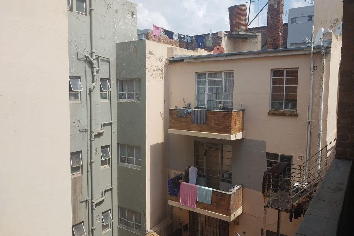 Exterior view of an apartment building rear facade showing multiple balconies, steel-framed windows, and visible signs of weathering and structural wear.
