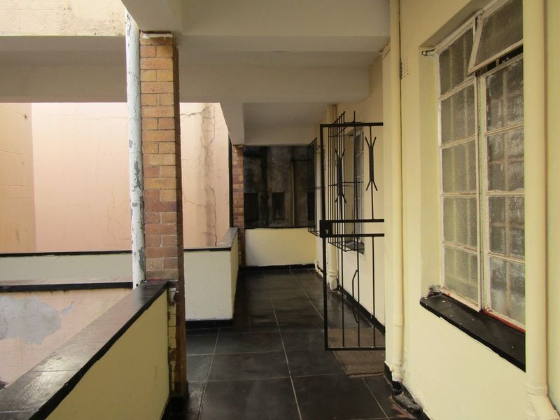 An outdoor balcony area featuring tiled flooring, a brick support pillar, and metal security gate, showing signs of wear and weathering on the exterior walls.