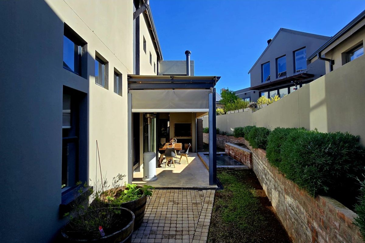 A well-maintained rear exterior featuring a covered patio with a built-in braai, paved walkway, and tiered garden beds with brick retaining walls.