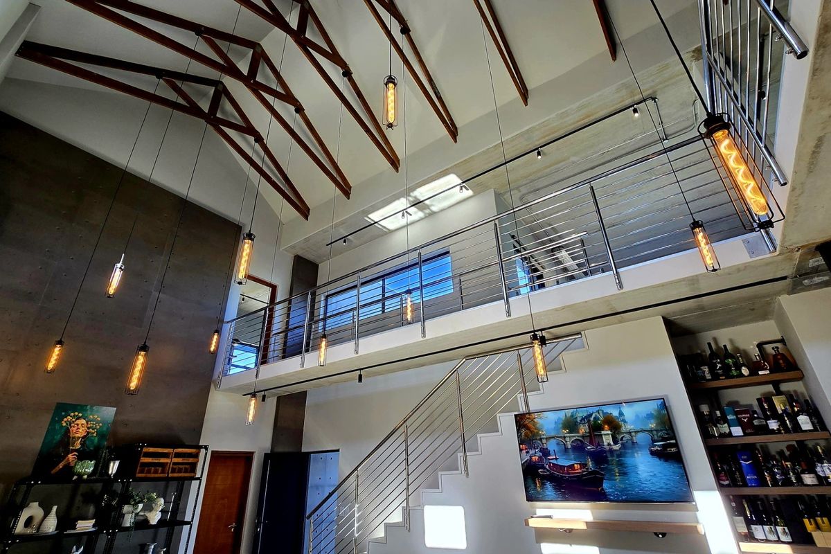 Spacious double-volume living area featuring exposed timber trusses, a mezzanine level with stainless steel balustrades, and modern industrial-style pendant lighting.