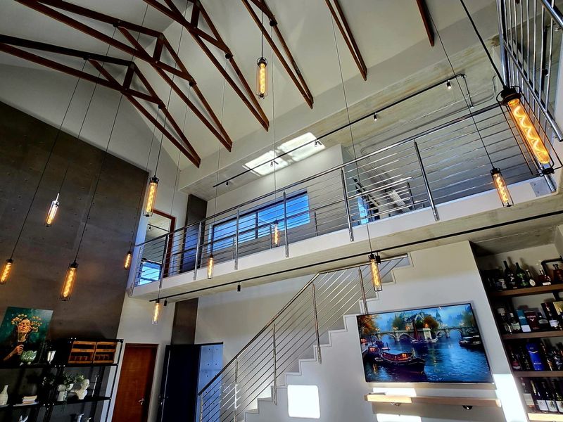 Spacious double-volume living area featuring exposed timber trusses, a mezzanine level with stainless steel balustrades, and modern industrial-style pendant lighting.