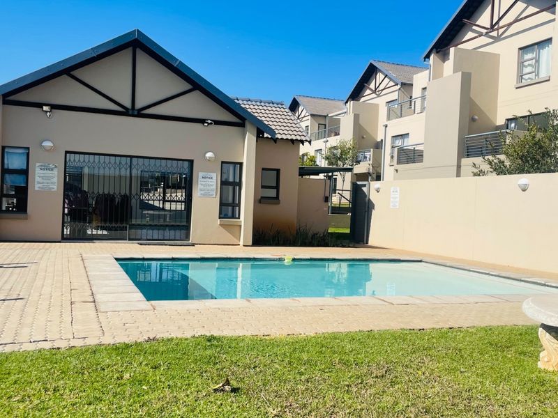 Communal swimming pool area featuring paved surround, manicured lawn, and secure perimeter walling within a well-maintained apartment complex.