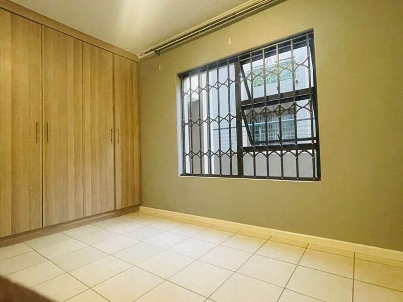 A clean, empty bedroom featuring neutral tiled flooring, built-in wooden cupboards, and a window fitted with a security gate.