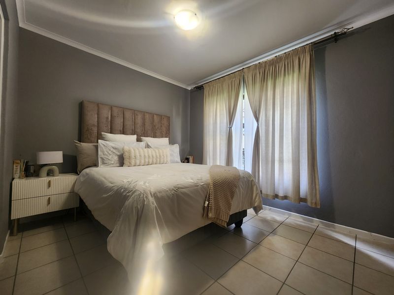 A well-maintained bedroom featuring neutral tiled flooring, grey painted walls, and large windows dressed with floor-to-ceiling curtains.