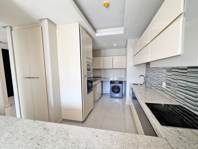 Modern kitchen featuring sleek cream cabinetry, granite countertops, integrated appliances, and a stylish mosaic tile backsplash with neutral tiled flooring.