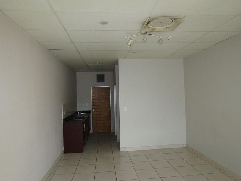 An empty open-plan living area featuring a compact kitchenette with built-in cabinetry, tiled flooring, and a suspended ceiling.