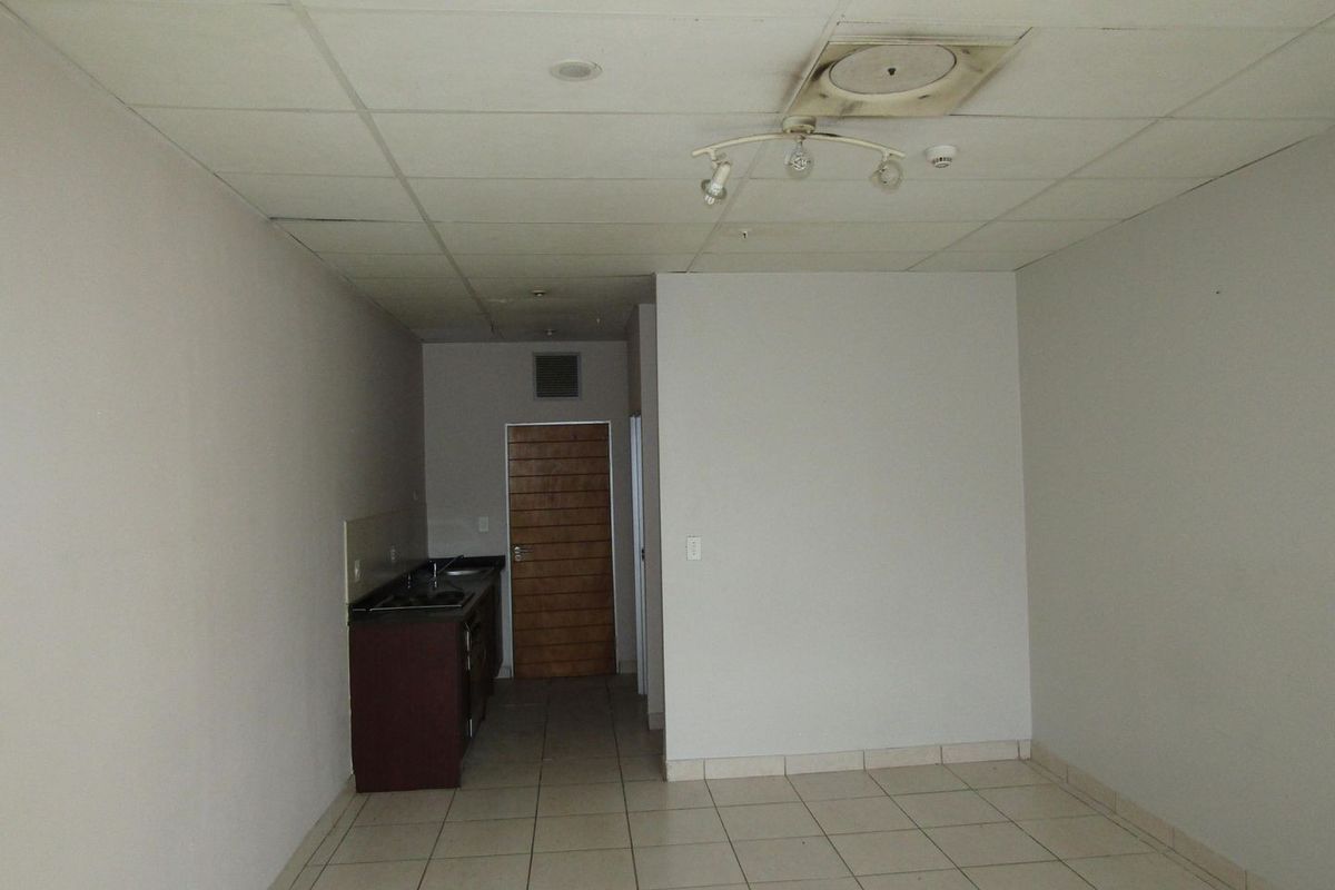 An empty open-plan living area featuring a compact kitchenette with built-in cabinetry, tiled flooring, and a suspended ceiling.