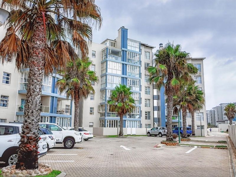 Exterior view of a multi-story apartment complex featuring a paved parking area, landscaped palm trees, and enclosed balcony structures.