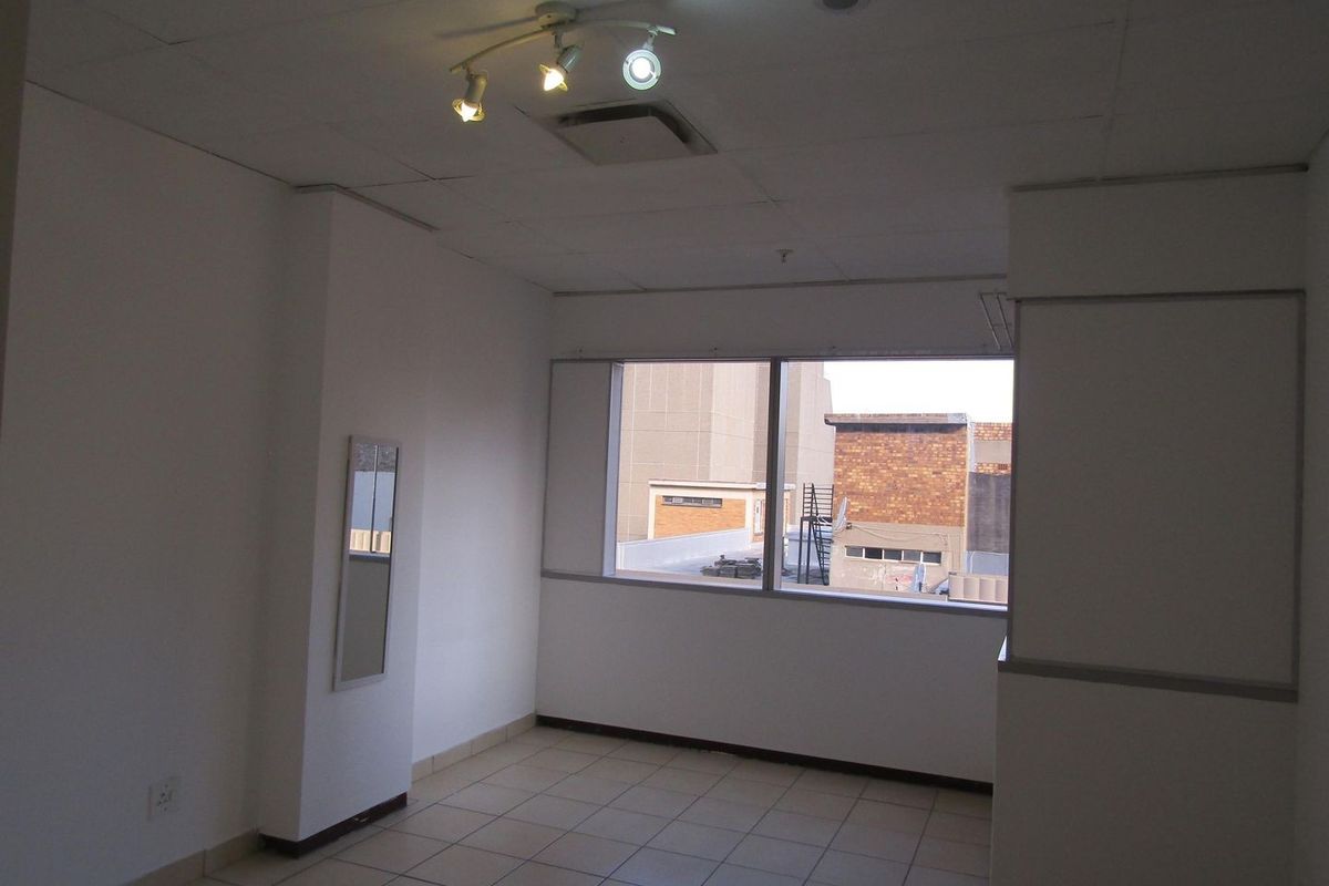 An empty, neutrally painted room featuring tiled flooring, a drop ceiling with track lighting, and a large window providing natural light.