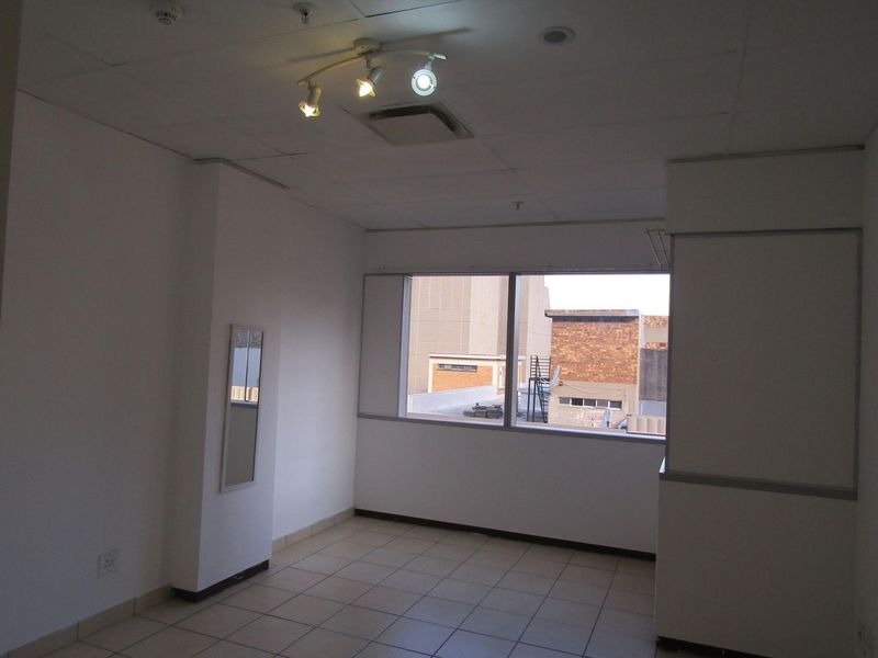 An empty, neutrally painted room featuring tiled flooring, a drop ceiling with track lighting, and a large window providing natural light.