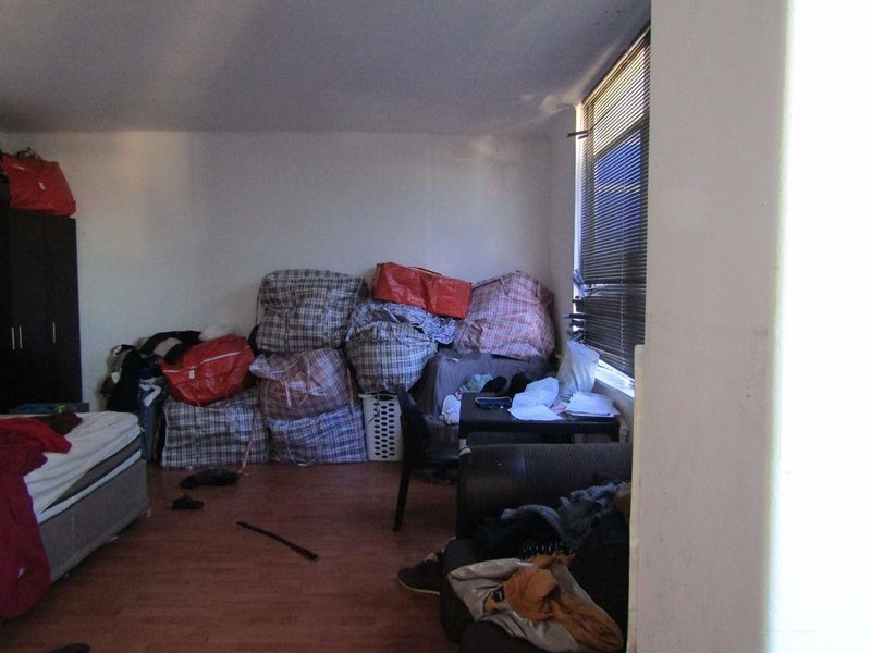 A bedroom featuring laminate flooring and window blinds, currently cluttered with personal belongings.