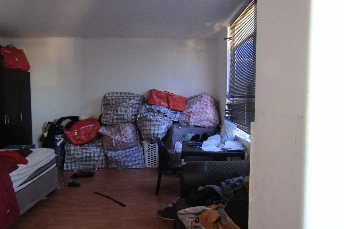A bedroom featuring laminate flooring and window blinds, currently cluttered with personal belongings.