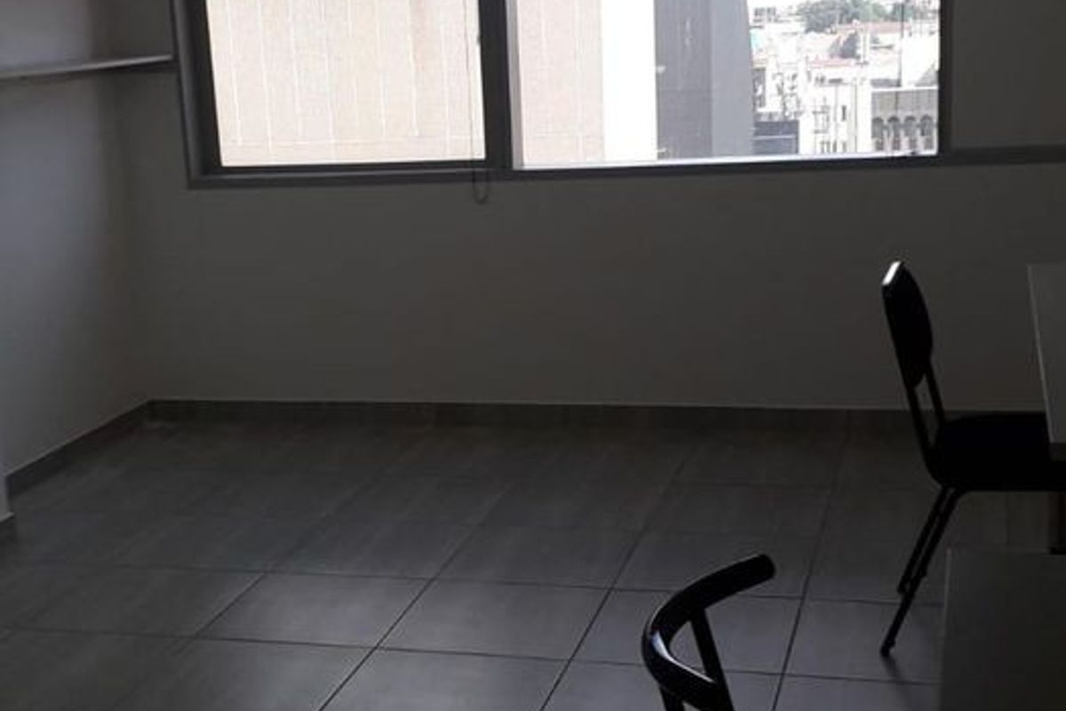 A sparsely furnished living area featuring large windows with city views, tiled flooring, and built-in shelving, offering a bright and functional space.