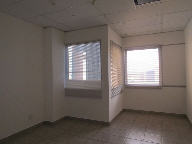 An empty office space featuring tiled flooring, a suspended ceiling, and windows fitted with venetian blinds.