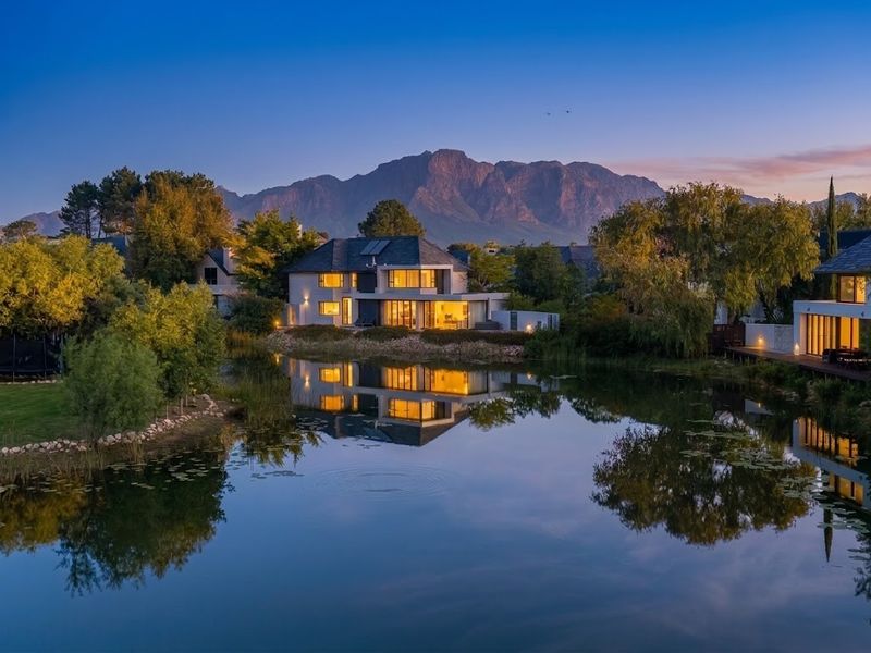 A stunning rear view of a modern residence at dusk, featuring expansive glass doors, landscaped grounds, and a tranquil water feature against mountain views.