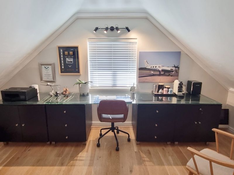 A well-lit attic office space featuring timber flooring, sloped ceilings, and built-in desk cabinetry under a central window.