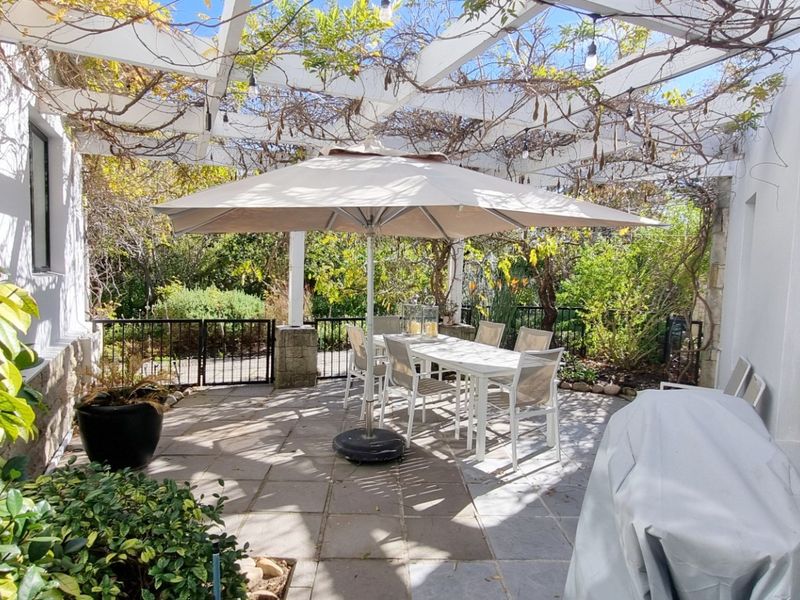 A charming outdoor patio area featuring a white pergola structure, stone paving, and established garden surroundings, perfect for alfresco dining and relaxation.