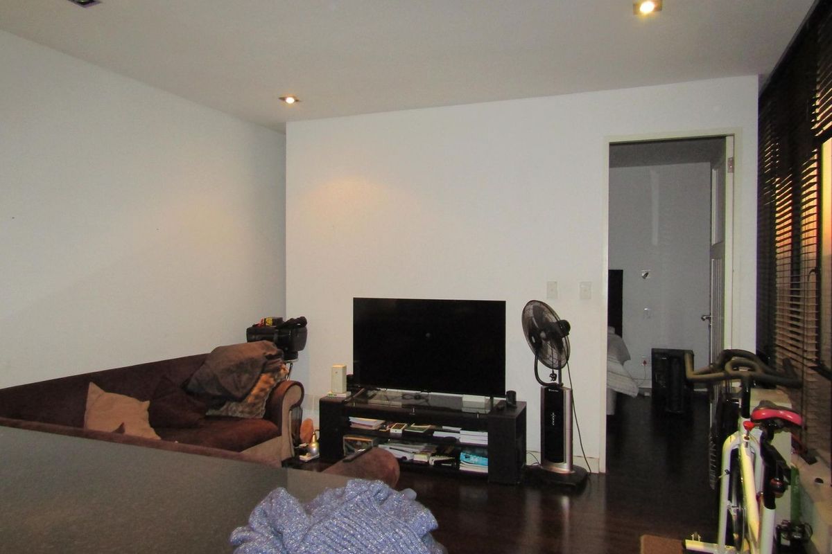 A living room featuring dark timber flooring, recessed downlighting, and venetian blinds, providing a functional space with direct access to an adjoining room.