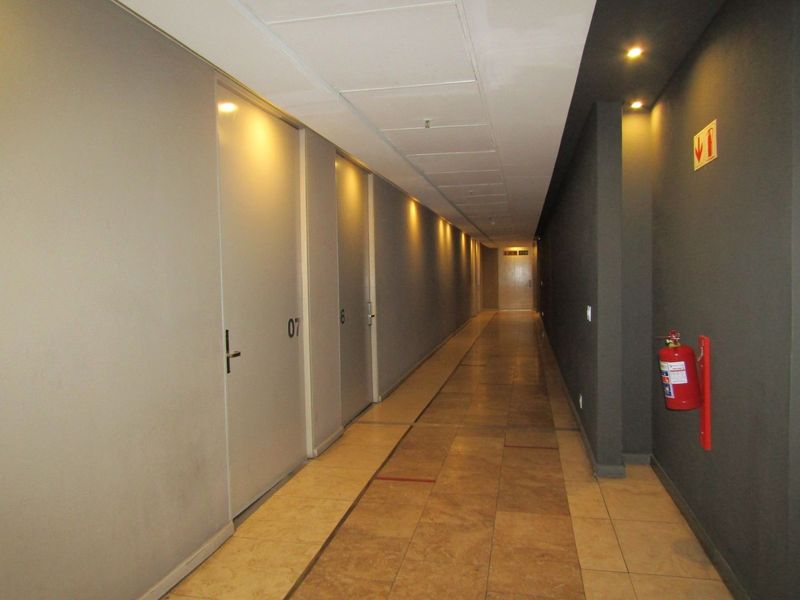 A well-lit apartment building hallway featuring tiled flooring, recessed ceiling lighting, and multiple unit access doors with integrated fire safety equipment.