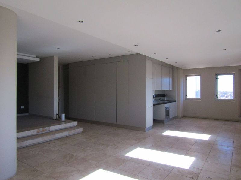 Spacious open-plan living area featuring neutral stone tile flooring, recessed lighting, and a compact built-in kitchen unit with integrated oven.