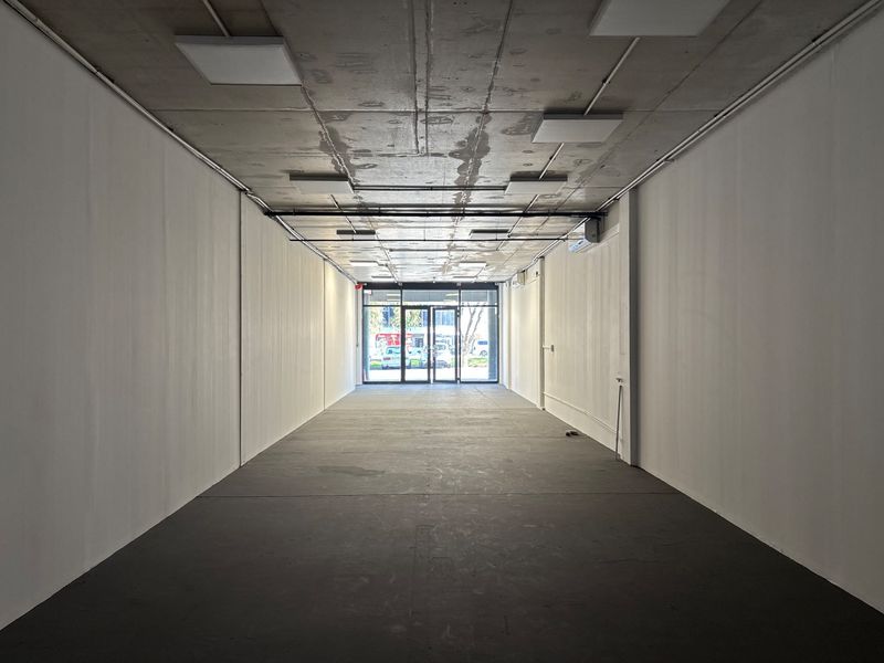 An empty, open-plan retail space featuring an exposed concrete ceiling, modern lighting, and a glass storefront entrance, suitable for various commercial uses.