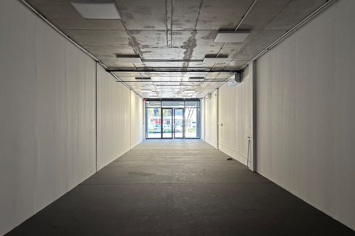 An empty, open-plan retail space featuring an exposed concrete ceiling, modern lighting, and a glass storefront entrance, suitable for various commercial uses.