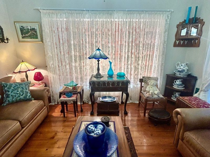 A cozy living room featuring polished timber flooring, high ceilings with decorative cornicing, and a large window dressed with sheer curtains.